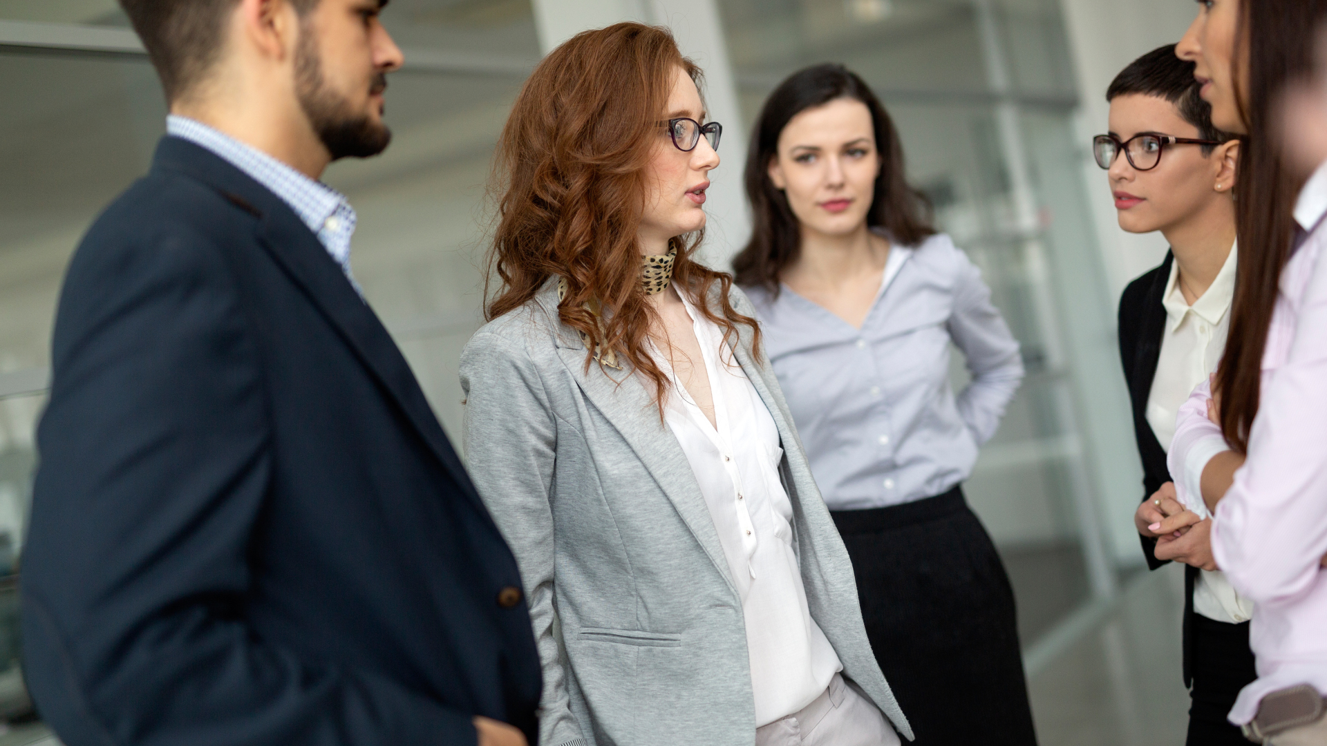 • Fünf Führungskräfte im Gespräch im Büroflur; eine Frau spricht, die anderen hören aufmerksam zu – Fokus auf Austausch und Sparring.