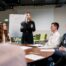 Führungskraft steht vor Flipchart und moderiert Teamgespräch am runden Tisch