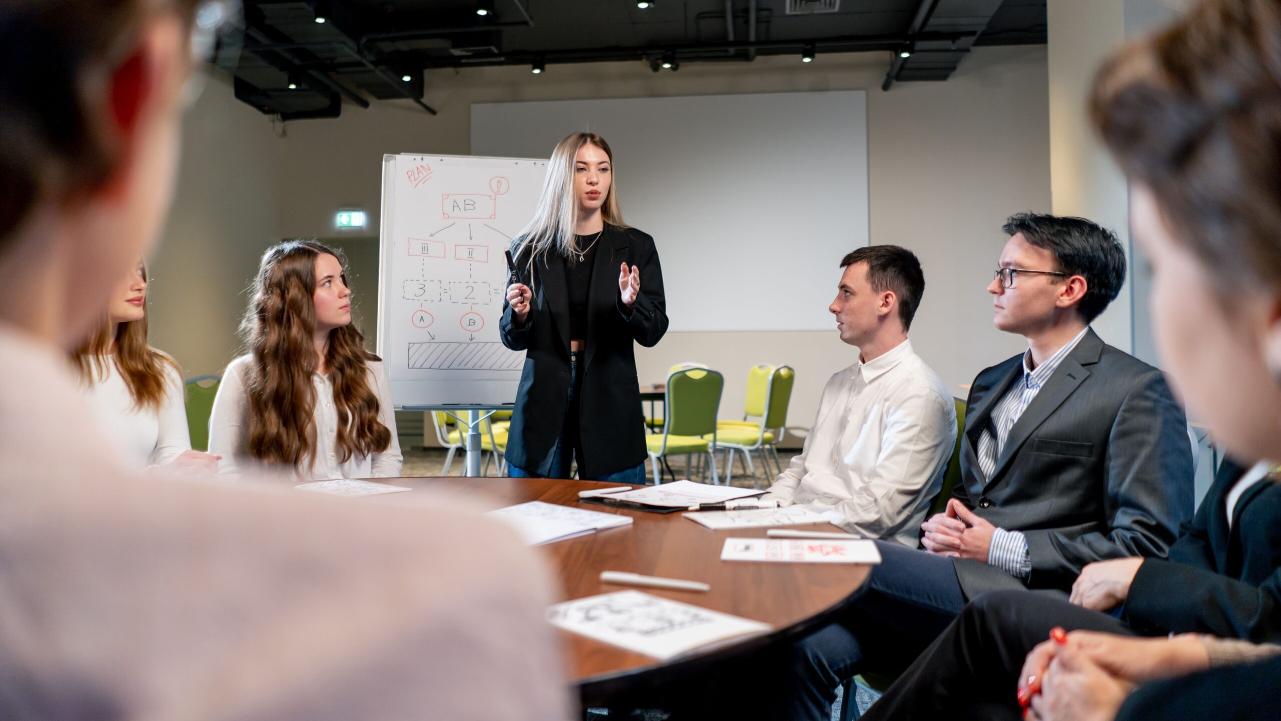 Führungskraft steht vor Flipchart und moderiert Teamgespräch am runden Tisch