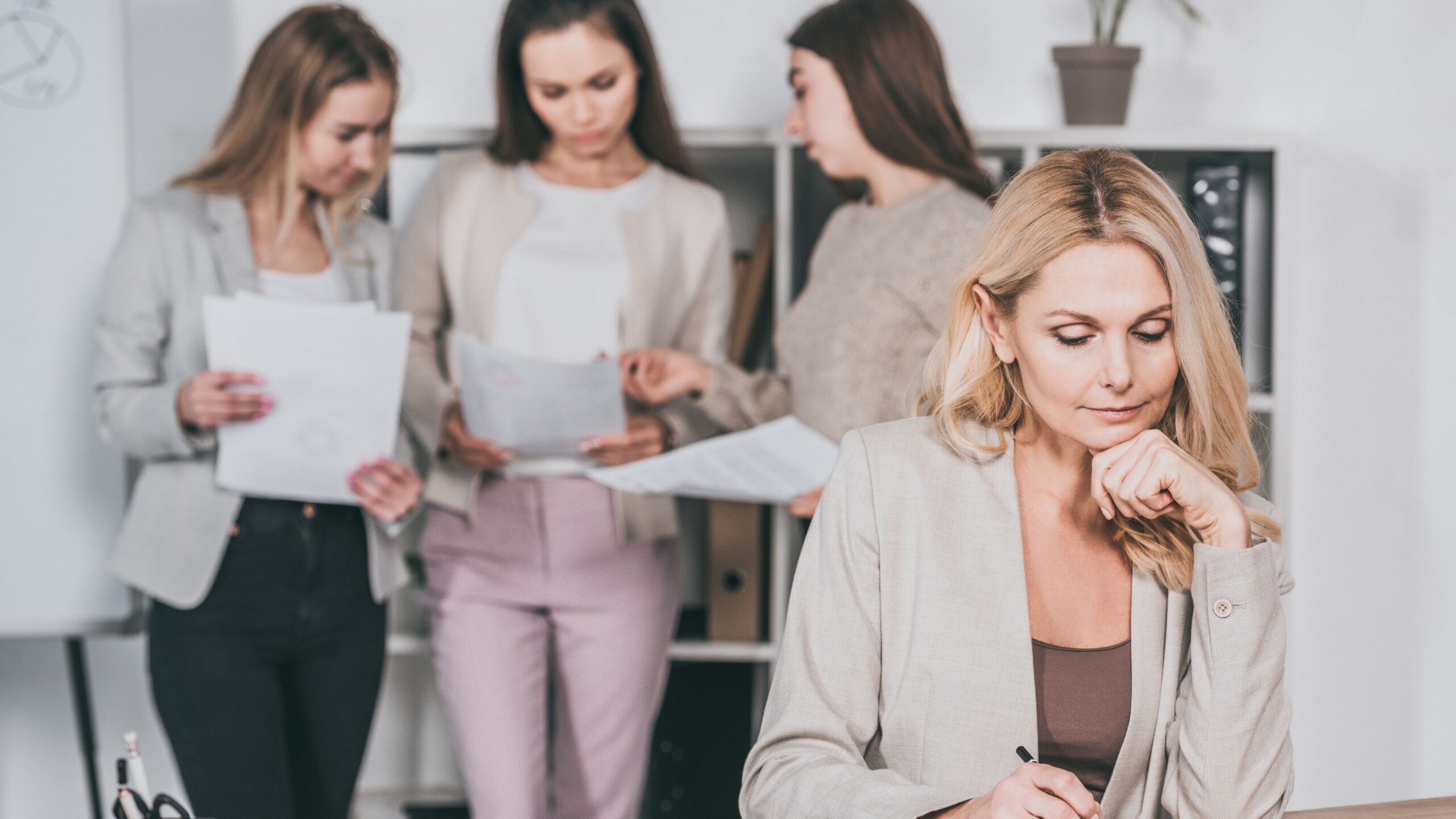 Führungskraft sitzt nachdenklich am Schreibtisch und schreibt, im Hintergrund unscharf drei Kolleginnen mit Dokumenten im Büro, Sinnbild für Vorbildwirkung in der Führung