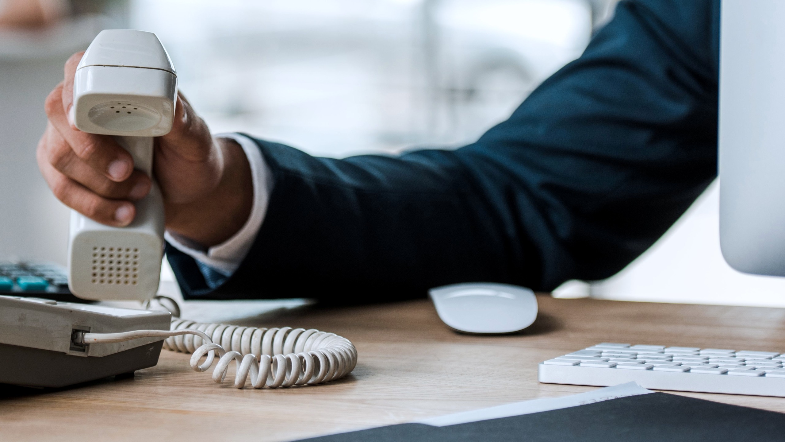 Ein Mann im Anzug hebt einen altmodischen Telefonhörer mit Wendelkabel vom Tisch, im Hintergrund unscharf Tastatur
